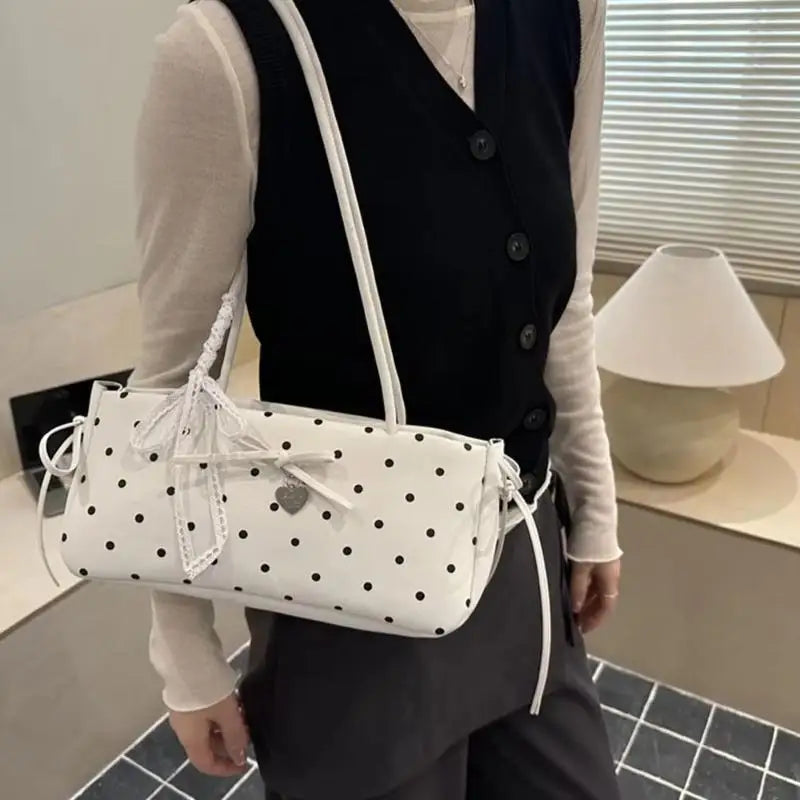 Eine Frau hält eine weiße Schultertasche mit schwarzen Polka Dots und einer weißen Schleife vor hellem Hintergrund.