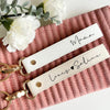 A white leather wristlet keychain with gold hardware and black script writing on a pink textured surface.