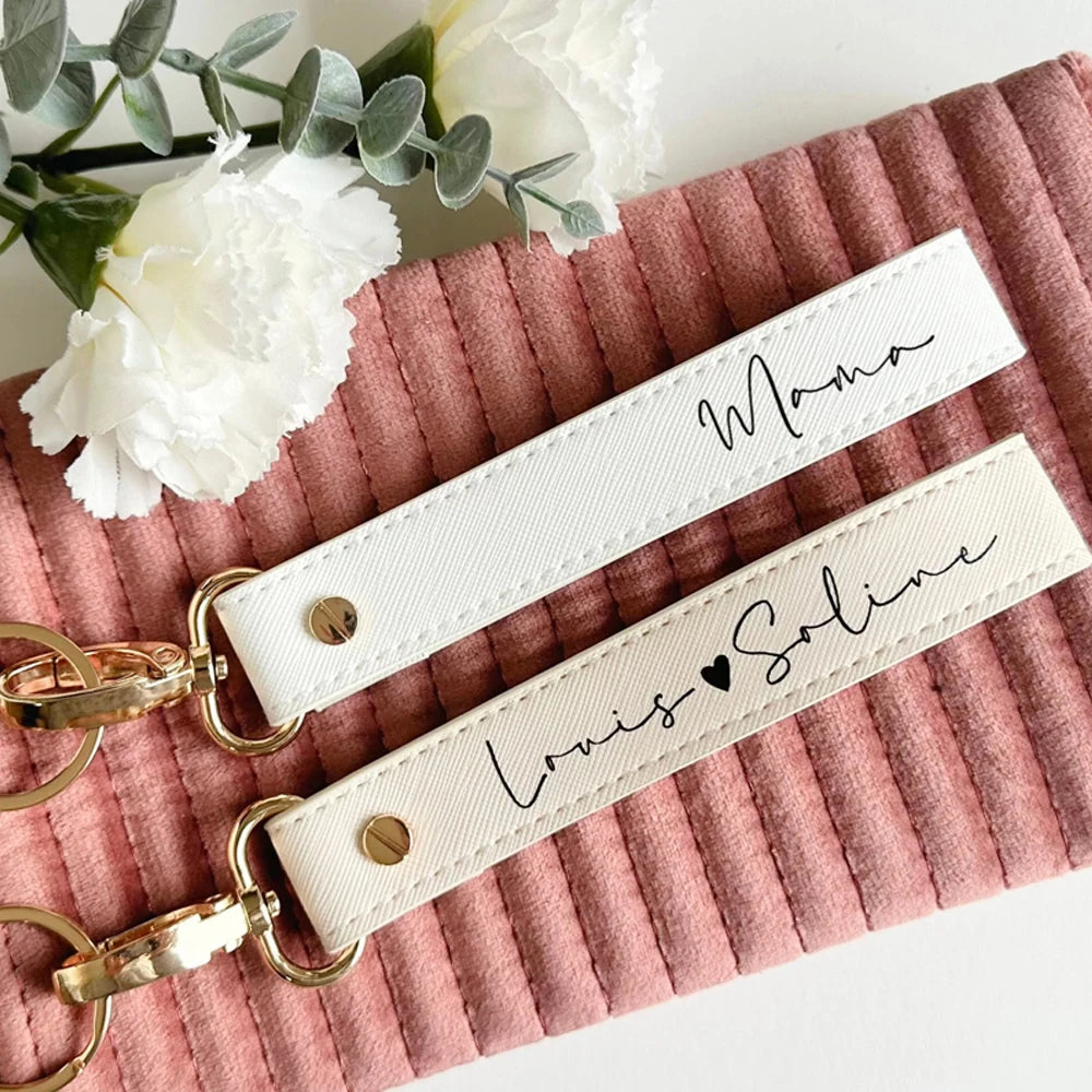 A white leather wristlet keychain with gold hardware and black script writing on a pink textured surface.