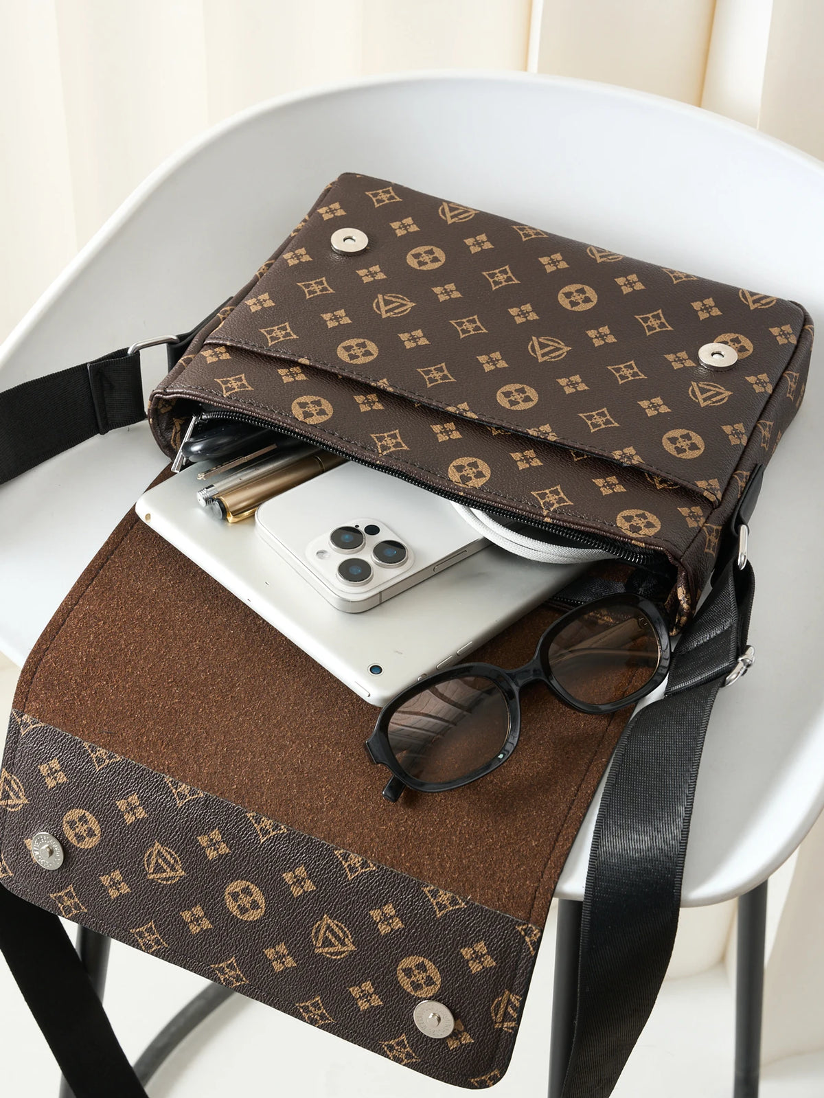 A brown patterned crossbody bag is placed on a white table next to a smartphone and sunglasses.