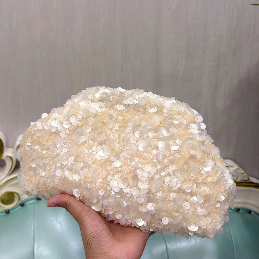 A hand holds a beige beaded chain shoulder clutch bag against a light background.