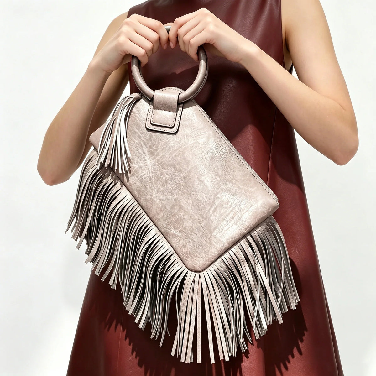 A woman holds a light pink fringed clutch bag with tassels against a dark red dress.