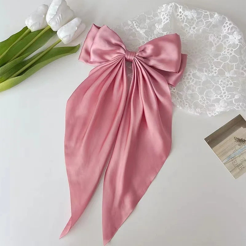 A pink silk bow hair clip is displayed next to white flowers and a lace fabric on a white surface.