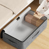 A gray foldable underbed storage bag is shown open on a wooden floor next to a bed with white bedding.