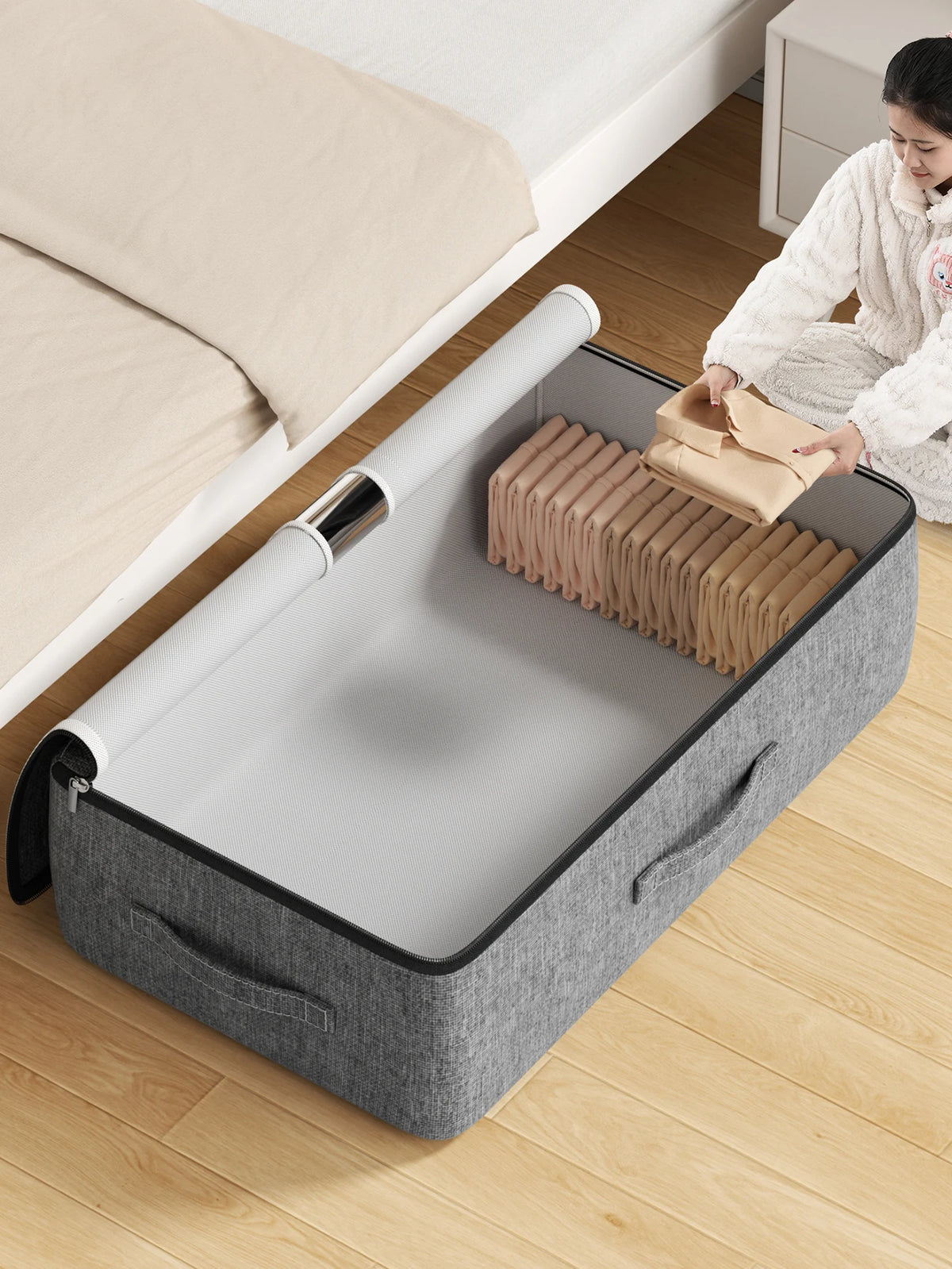 A gray foldable underbed storage bag is shown open on a wooden floor next to a bed with white bedding.