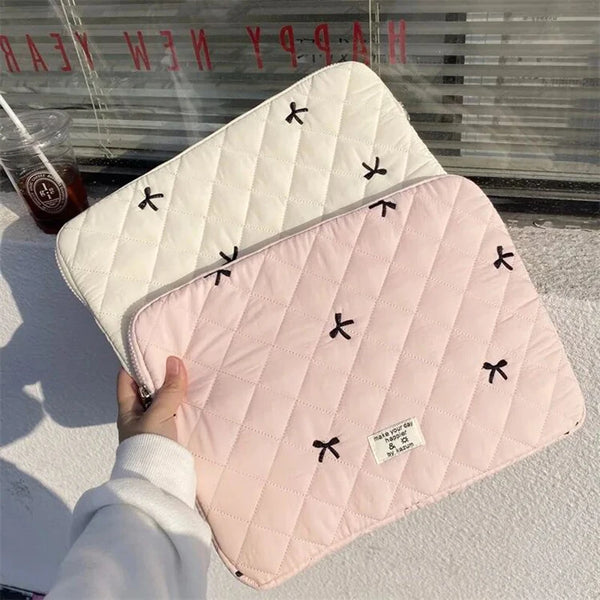 Housse de laptop ajustée en quilted rose avec broderie de nœud noir, devant un fond de fenêtre.