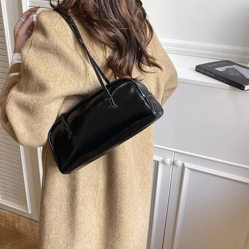 A woman wearing a beige coat holds a black glossy PU leather tote bag with dual straps.