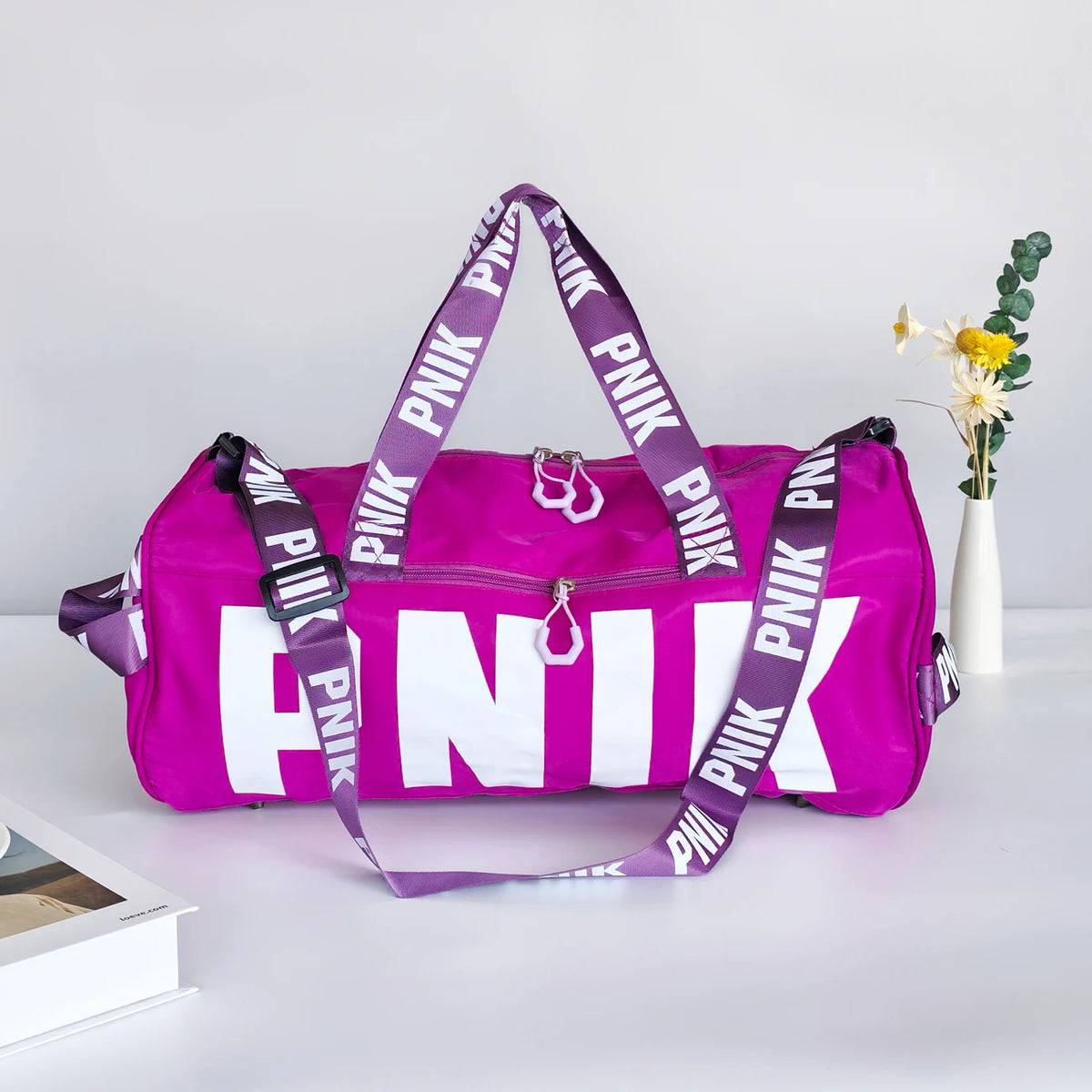 A bright purple Nike pink shoulder bag with white text is displayed on a white surface next to a small vase of yellow flowers