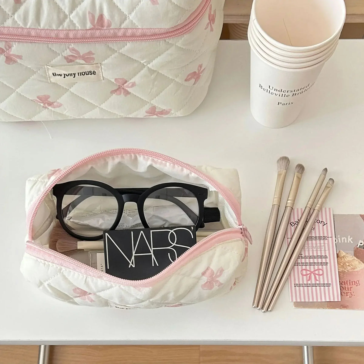 Sac de rangement cosmétique blanc avec motif nœud rose, à côté d'une tasse rose et de pinceaux de maquillage.