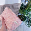 Una bolsa de almacenamiento con cremallera rosa de lona, sostenida contra un fondo de plantas tropicales verdes.