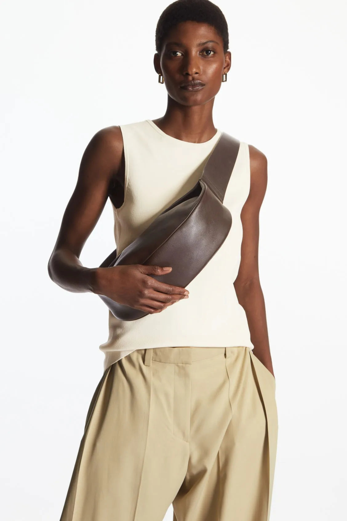 Modelo vistiendo un top sin mangas color crema y pantalones beige, sosteniendo un bolso cruz de cuero marrón.