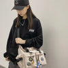 A young woman wearing a black hoodie and a black cap holds a beige nylon crossbody bag with various charms.