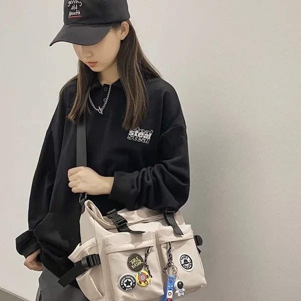 A young woman wearing a black hoodie and a black cap holds a beige nylon crossbody bag with various charms.