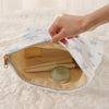 A hand is opening a light beige cosmetic bag with a white and blue starfish pattern, revealing a round jar inside.