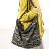 A person is holding a canvas camouflage shoulder bag with an adjustable strap against a white background.