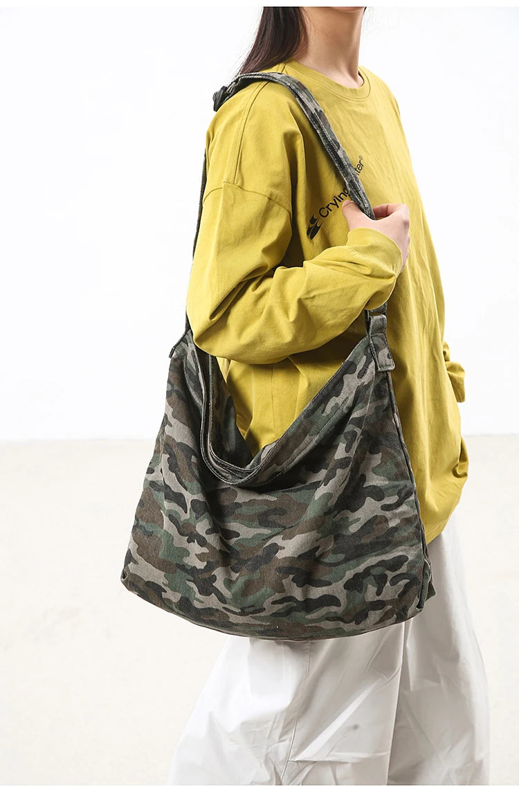A person is holding a canvas camouflage shoulder bag with an adjustable strap against a white background.