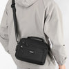 A man wearing a gray waterproof jacket is carrying a black compact crossbody messenger bag.