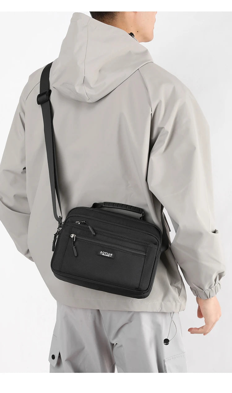 A man wearing a gray waterproof jacket is carrying a black compact crossbody messenger bag.