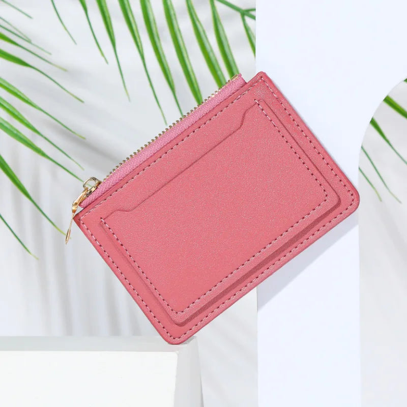 A pink PU leather mini card holder wallet with a zipper is shown against a white background with green leaves.