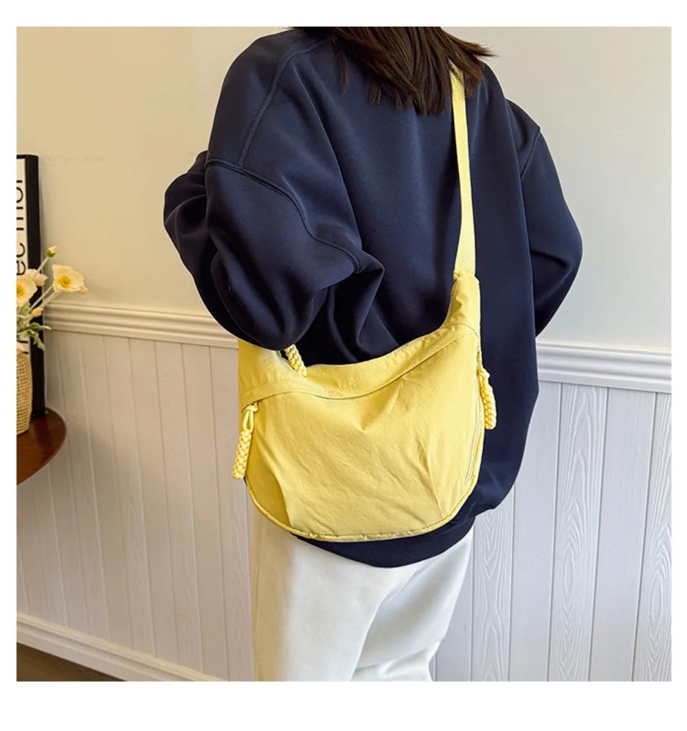 A yellow canvas crossbody bag is shown hanging from a navy blue sweatshirt, next to a white paneled wall.