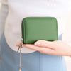 A hand holds a green accordion-style wallet with a zipper and chain attached, against a light background.