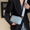 A woman holds a light blue vegan PU crossbody bag with a patterned strap against a black blazer.