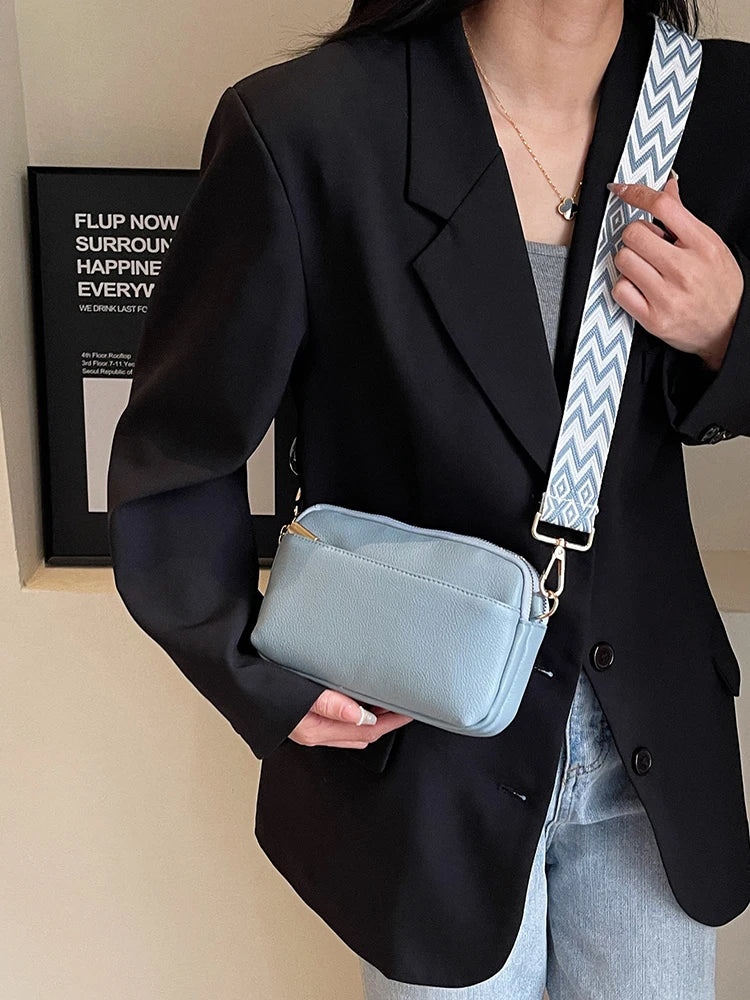 A woman holds a light blue vegan PU crossbody bag with a patterned strap against a black blazer.