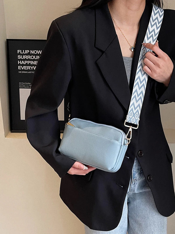 A woman holds a light blue vegan PU crossbody bag with a patterned strap against a black blazer.