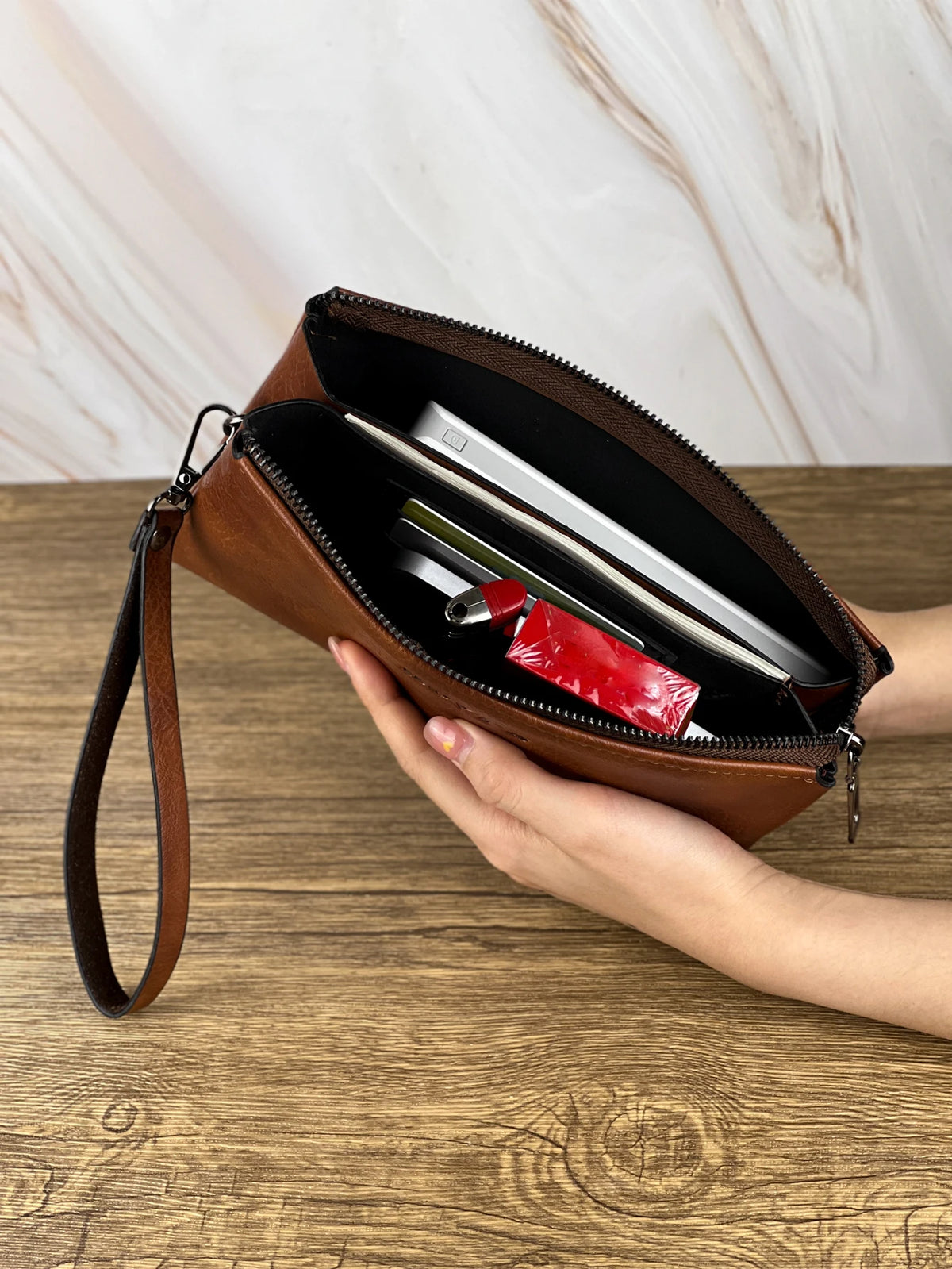 Une main tient un porte-monnaie clutch en cuir marron à fermeture éclair ouverte, montrant une carte argentée et un objet rou