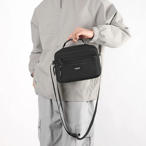 A man wearing a gray jacket is holding a black compact waterproof crossbody messenger bag.