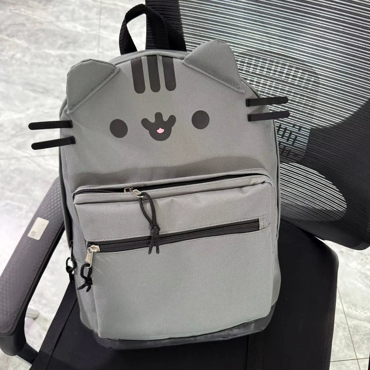 A gray Pusheen cat canvas backpack with black whiskers and eyes resting on a black chair.