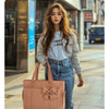 A young woman with long brown hair wearing a light blue sweatshirt and jeans is walking while carrying a large pink tote bag.