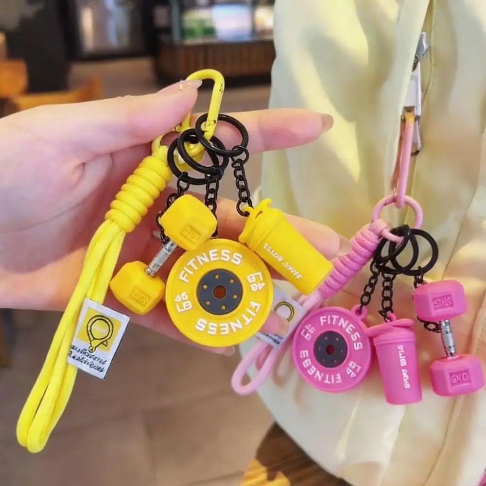 A hand holds a yellow fitness dumbbell keychain charm with black and pink components.