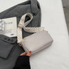 A light gray vegan PU crossbody bag with a beige and white patterned strap is being held by a person against a white backgrou