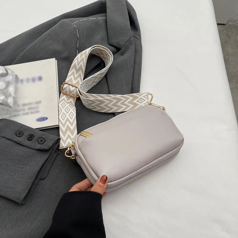 A light gray vegan PU crossbody bag with a beige and white patterned strap is being held by a person against a white backgrou