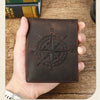 A dark brown leather wallet featuring an engraved compass design is held in a hand on a wooden surface.