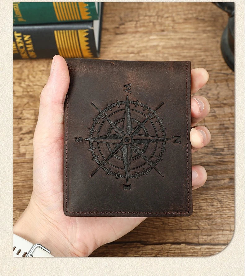 A dark brown leather wallet featuring an engraved compass design is held in a hand on a wooden surface.