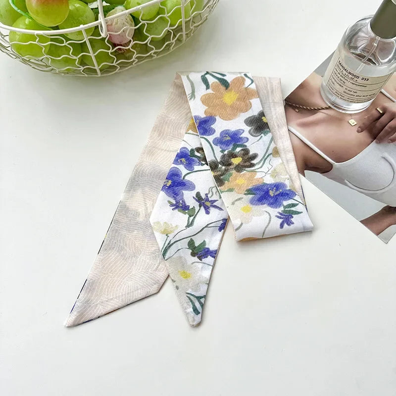 Un foulard de cheveux en soie beige avec des motifs floraux est exposé à côté d'une bouteille en verre et d'une main.