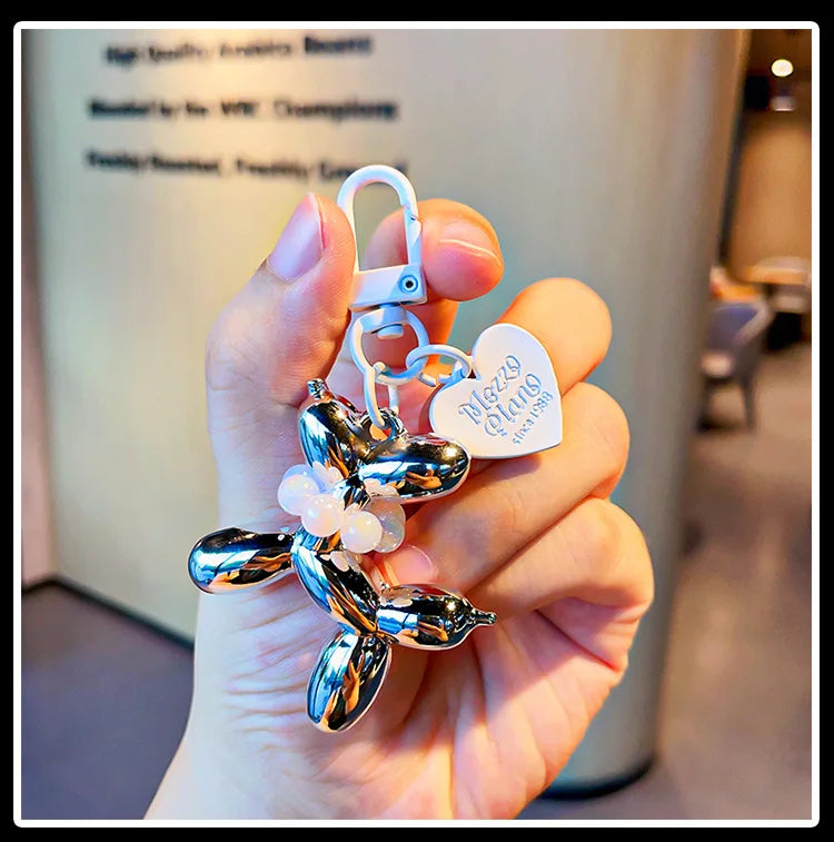 A hand holds a shiny metallic balloon dog keychain charm with a loop and a heart tag.