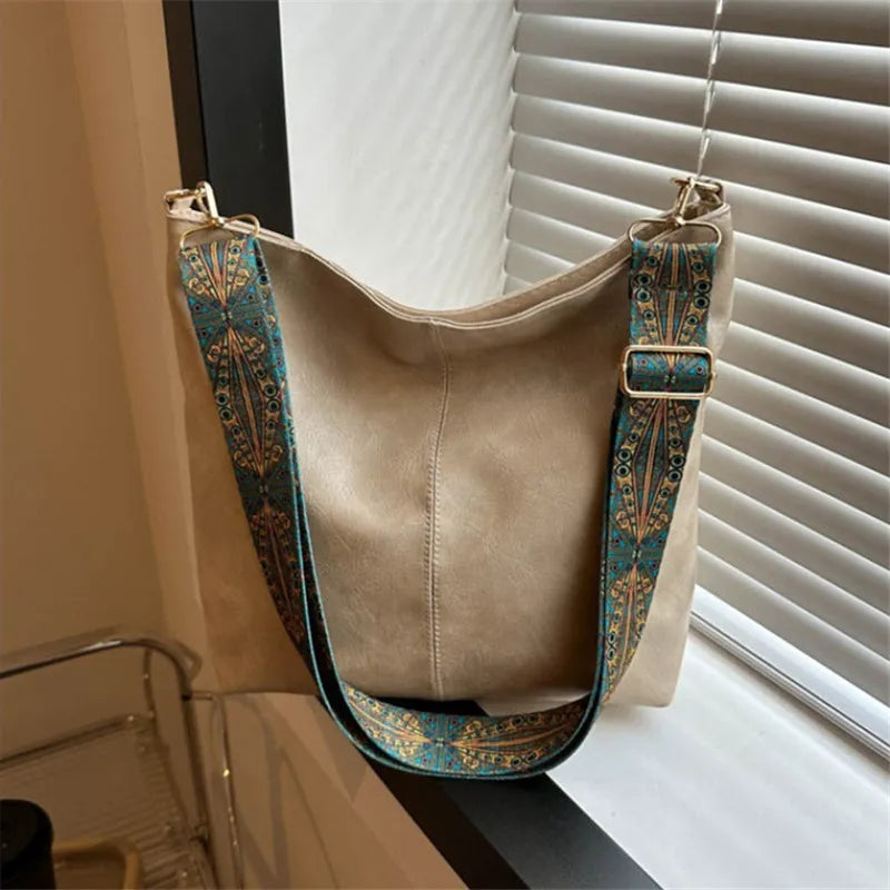 A tan shoulder bag with a wide strap featuring teal and gold embroidery, displayed next to a window blind.
