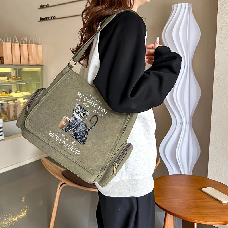 A person holds a olive green canvas messenger bag with a gray cat print featuring text on the front.