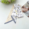 Une pile de foulards pour cheveux en soie élégants avec des motifs floraux blancs et gris sont exposés sur une surface blanch