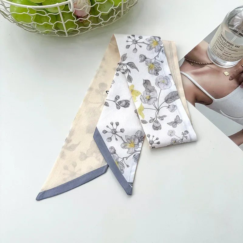 Une pile de foulards pour cheveux en soie élégants avec des motifs floraux blancs et gris sont exposés sur une surface blanch