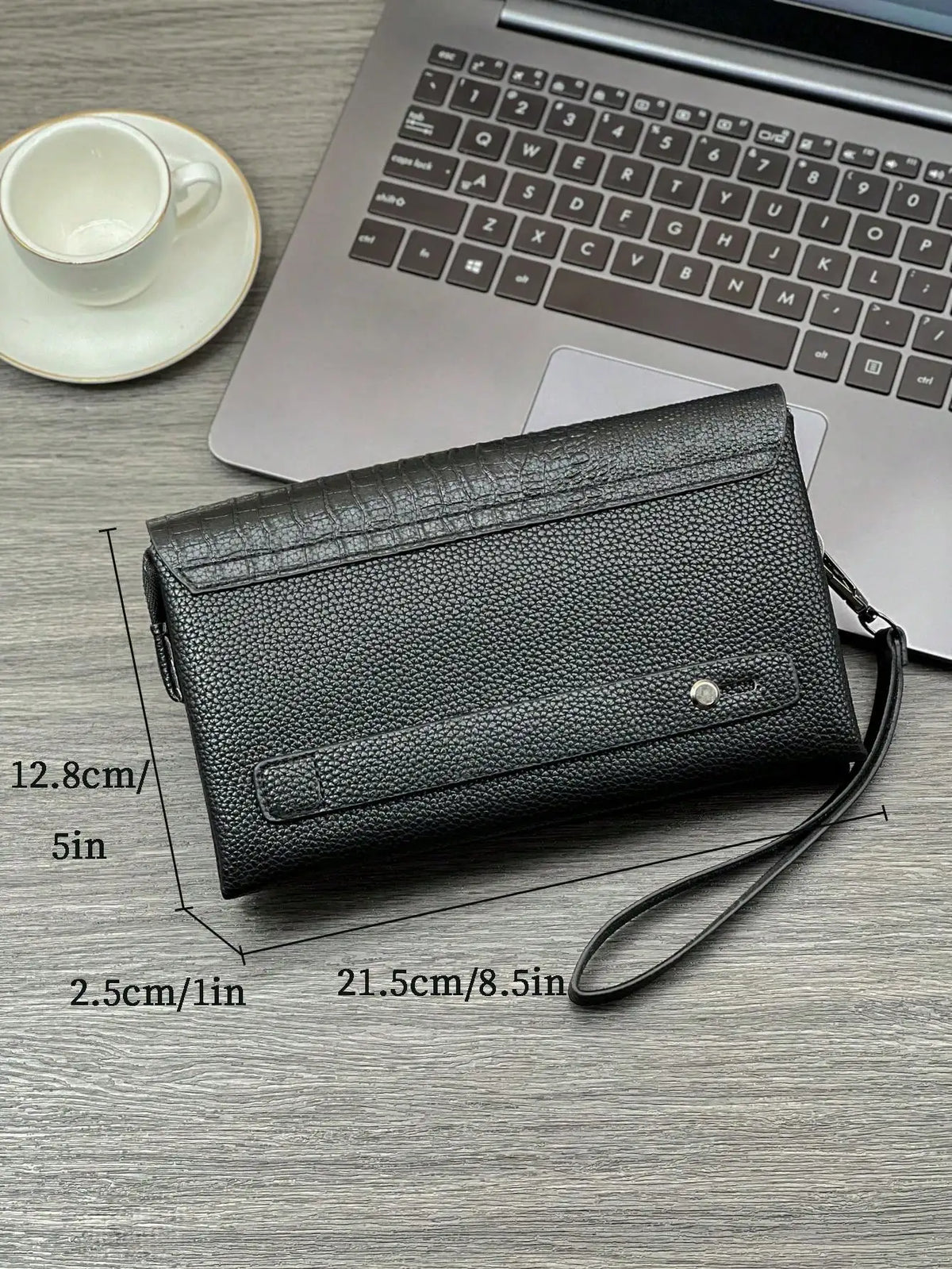A black crocodile pattern clutch bag lying on a wooden desk next to a silver laptop and a white cup.