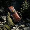 A hand wearing a green glove holds a brown leather knife sheath with a decorative pattern against a dark, rocky background.