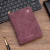 A deep plum colored slim passport wallet with gold accents resting on a wooden surface next to a laptop keyboard.