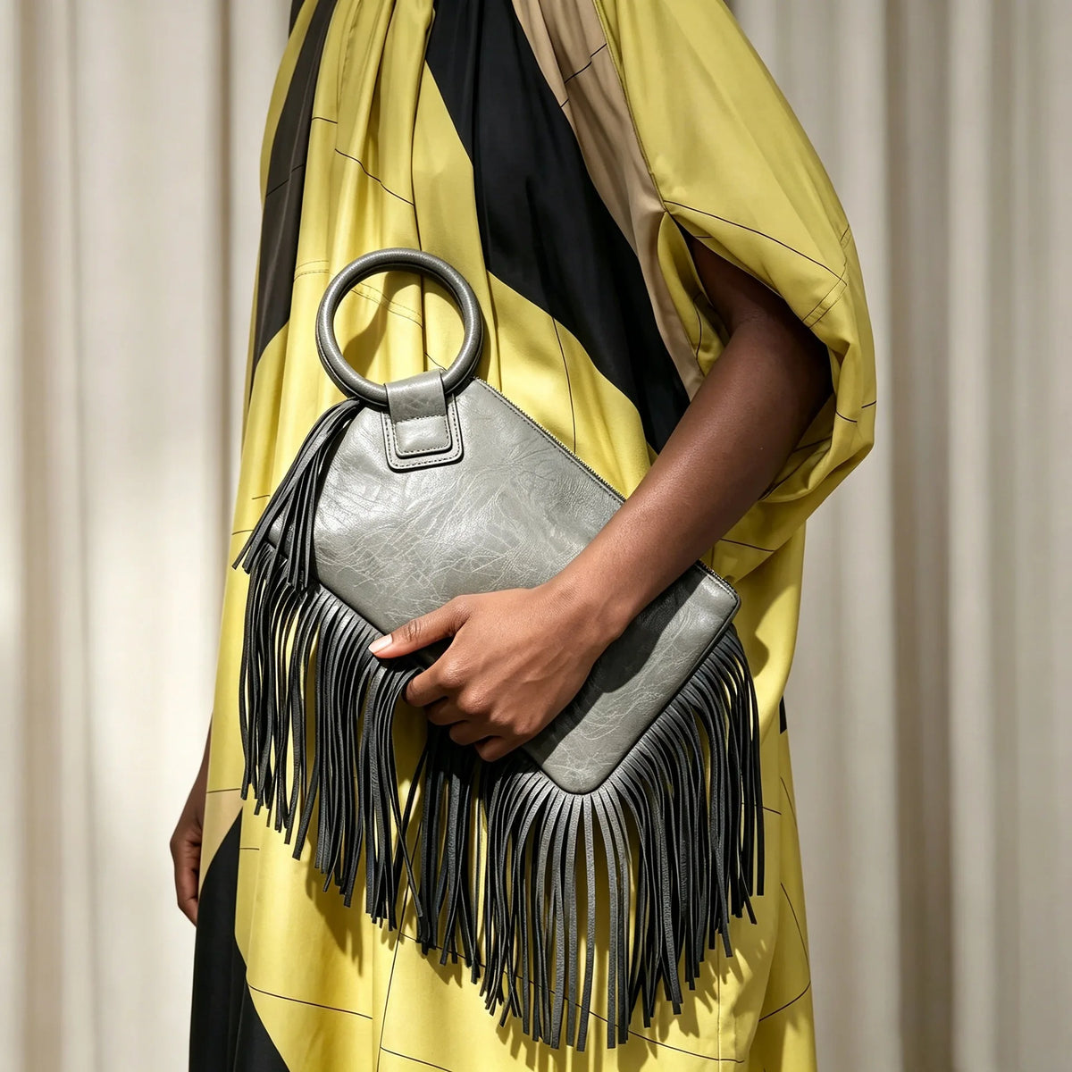 A hand holds a silver fringed clutch bag with black tassels against a yellow and black striped garment.
