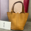 A tan genuine leather bucket bag with a wide handle is displayed against a blurred background of purple and pink fabric.