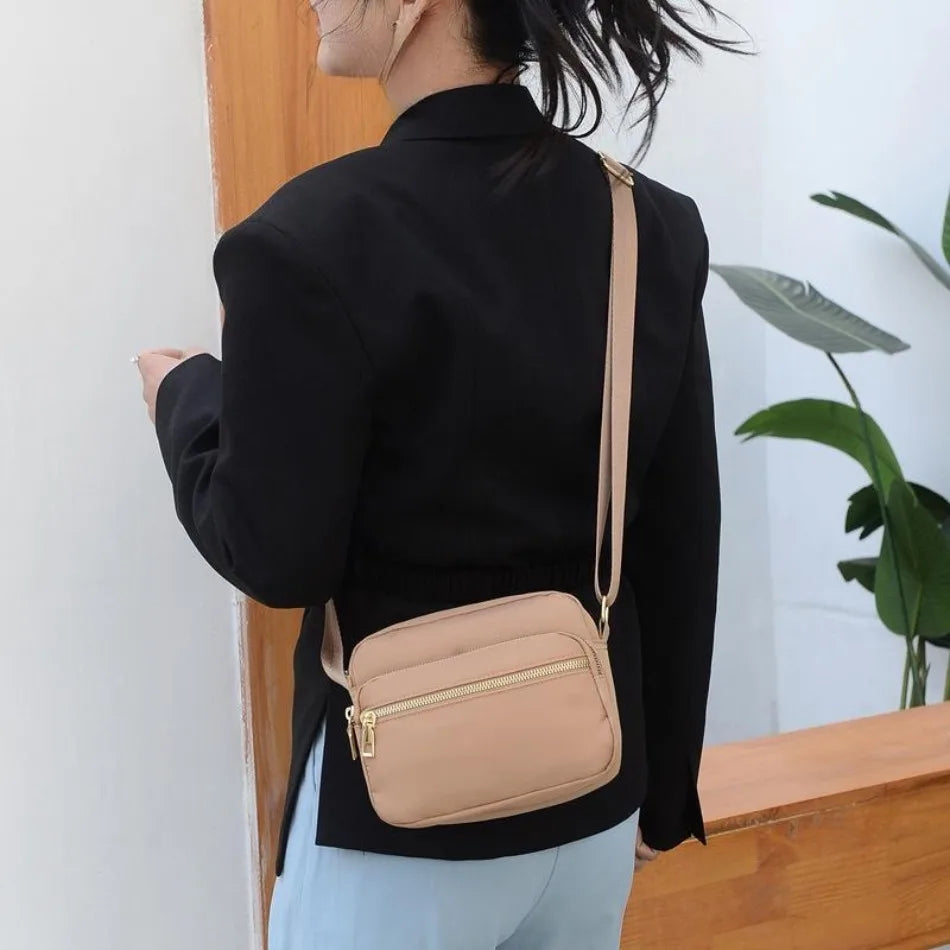 A woman wearing a black blazer and light blue pants holds a beige compact oxford fabric crossbody bag.