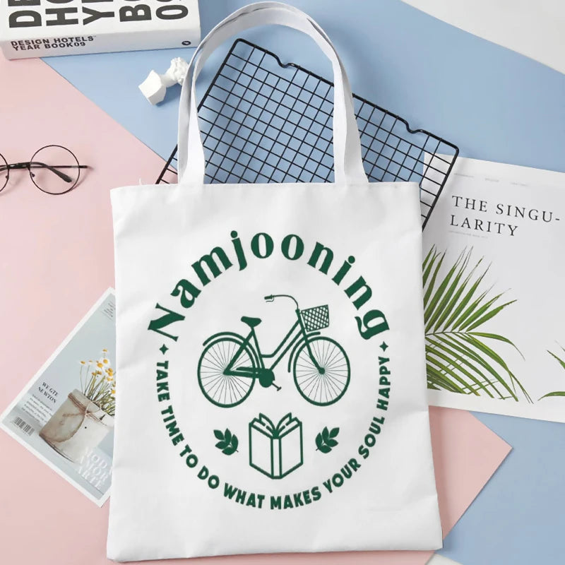 A white canvas tote bag featuring a green graphic design with the text Namjooning and a bicycle illustration.